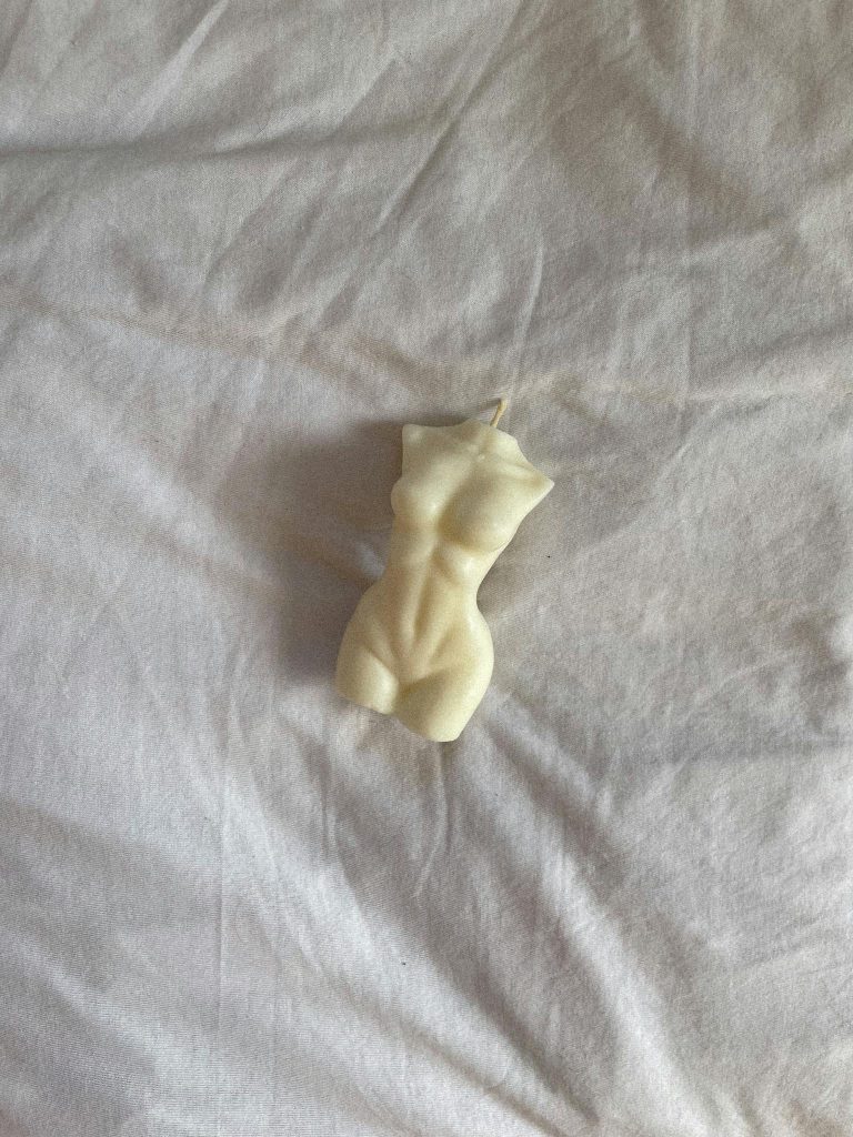 An artistic sculptural candle shaped like a human torso laying on a white sheet.